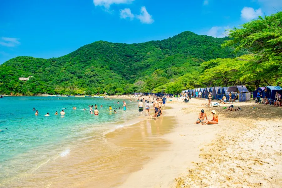 Bahía Concha: playa de arena con carpas azules, agua turquesa y una montaña verde al fondo.